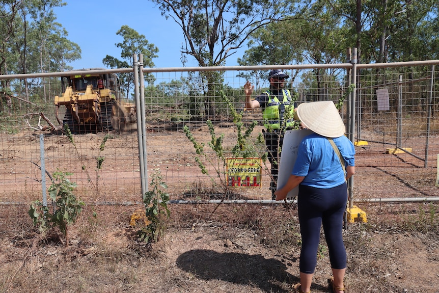 Lady wearing hat and holding sign stands before fence with a policeman and tractor behind it 