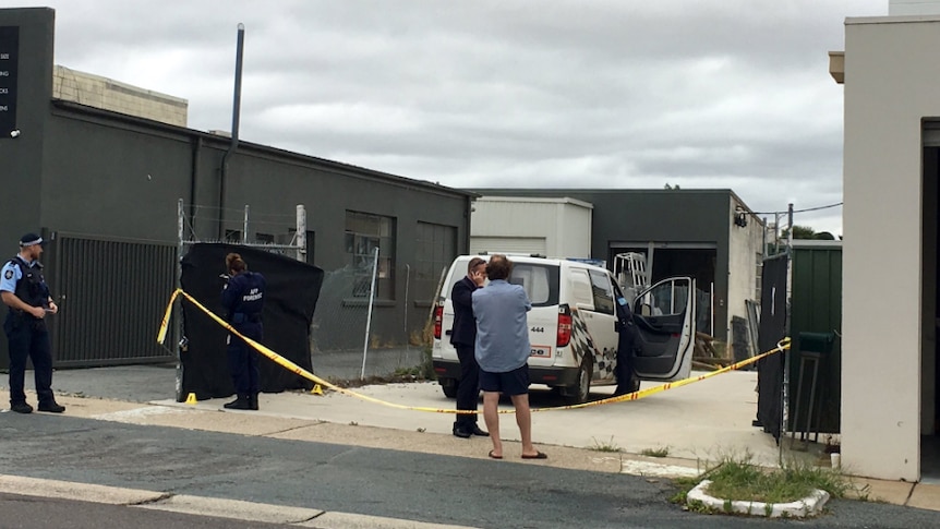 Police stand outside an industrial building, behind yellow police tape.