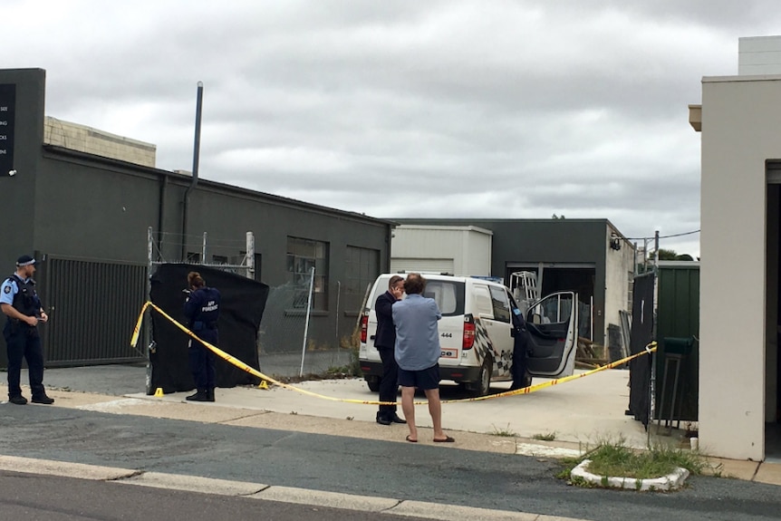 Police stand outside an industrial building, behind yellow police tape.