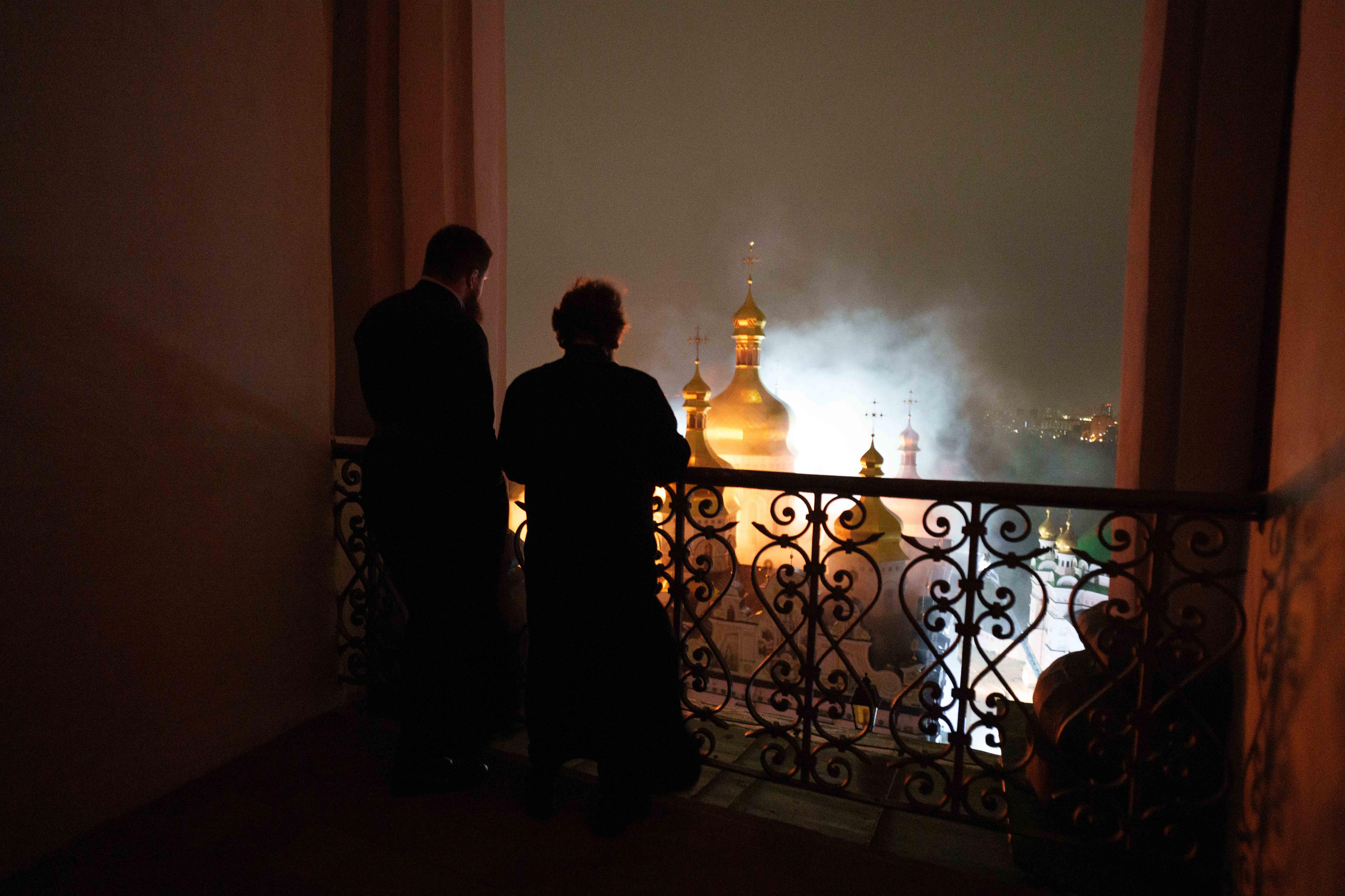 Silhouttes of two people standing at a window overlooking a cathedral up in flames. 