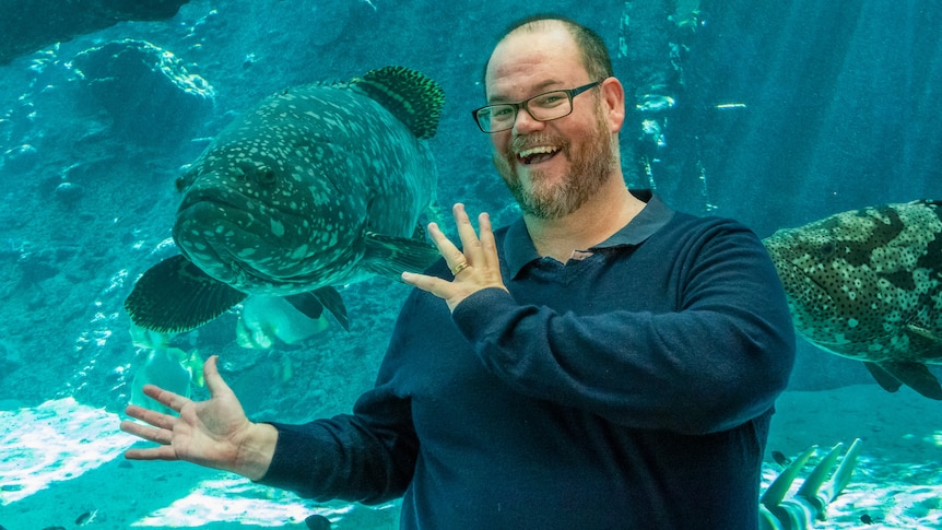 A man with a beard and glasses standing in front of an aquarium, with a large fish to his right.