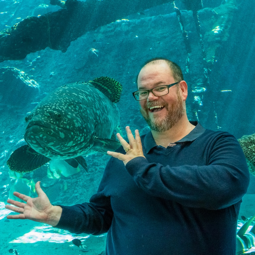 A man with a beard and glasses standing in front of an aquarium, with a large fish to his right.