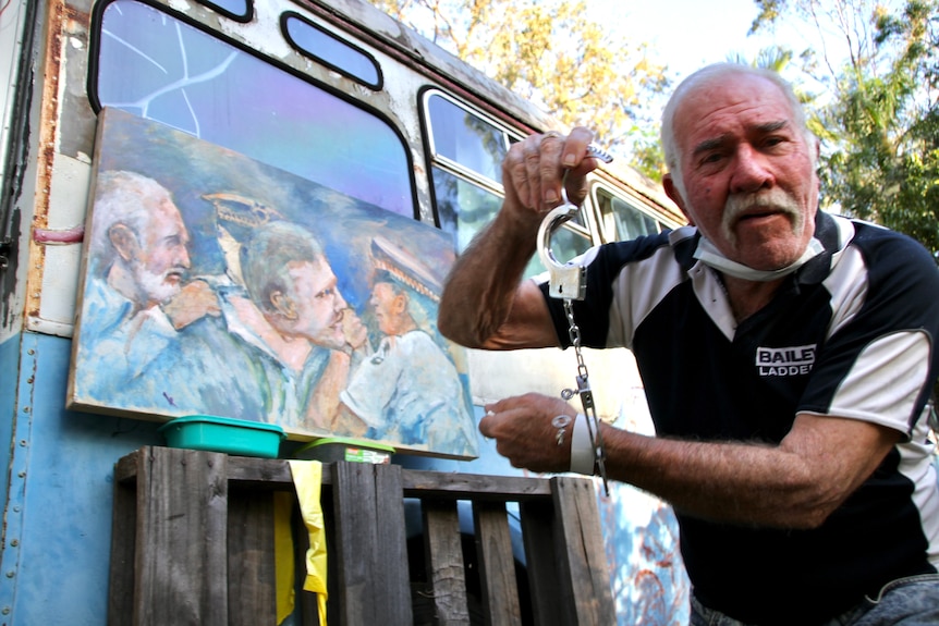 Grey haired man in handcuffs against a background of a painting 