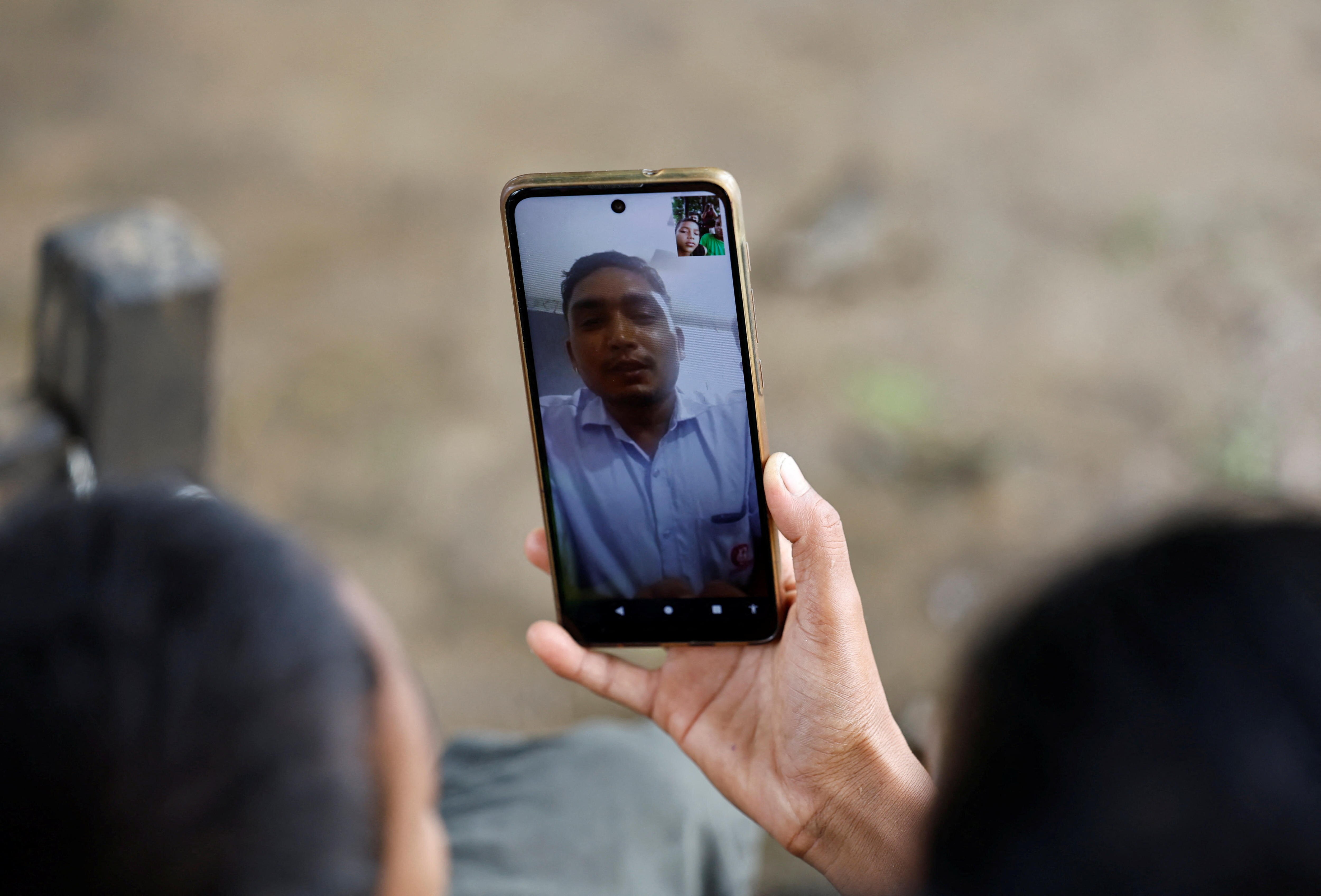 A hand holds a smartphone for a video call with a man in a blue collared shirt 
