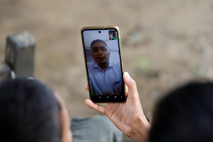 A hand holds a smartphone for a video call with a man in a blue collared shirt 