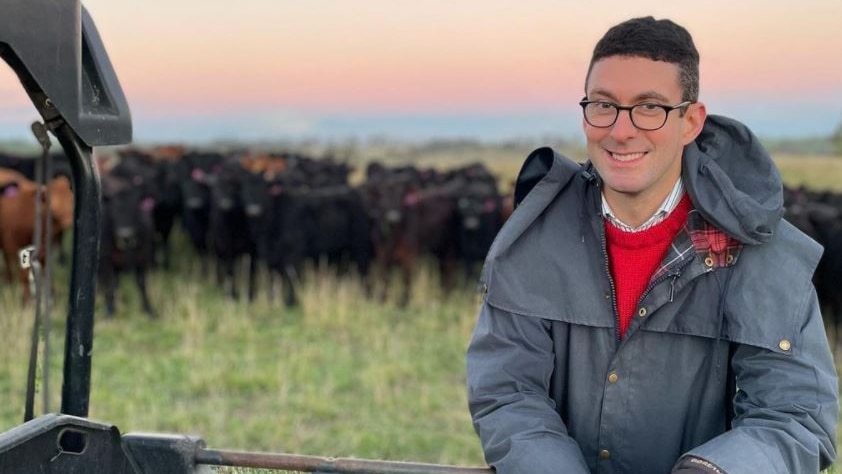 A man with glasses on a farm with cattle in the background