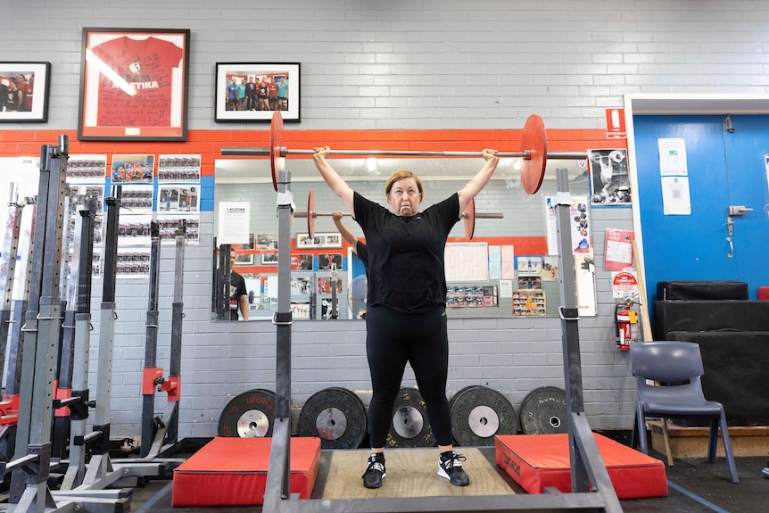 A middle-aged woman stands and lifts weights above her head.