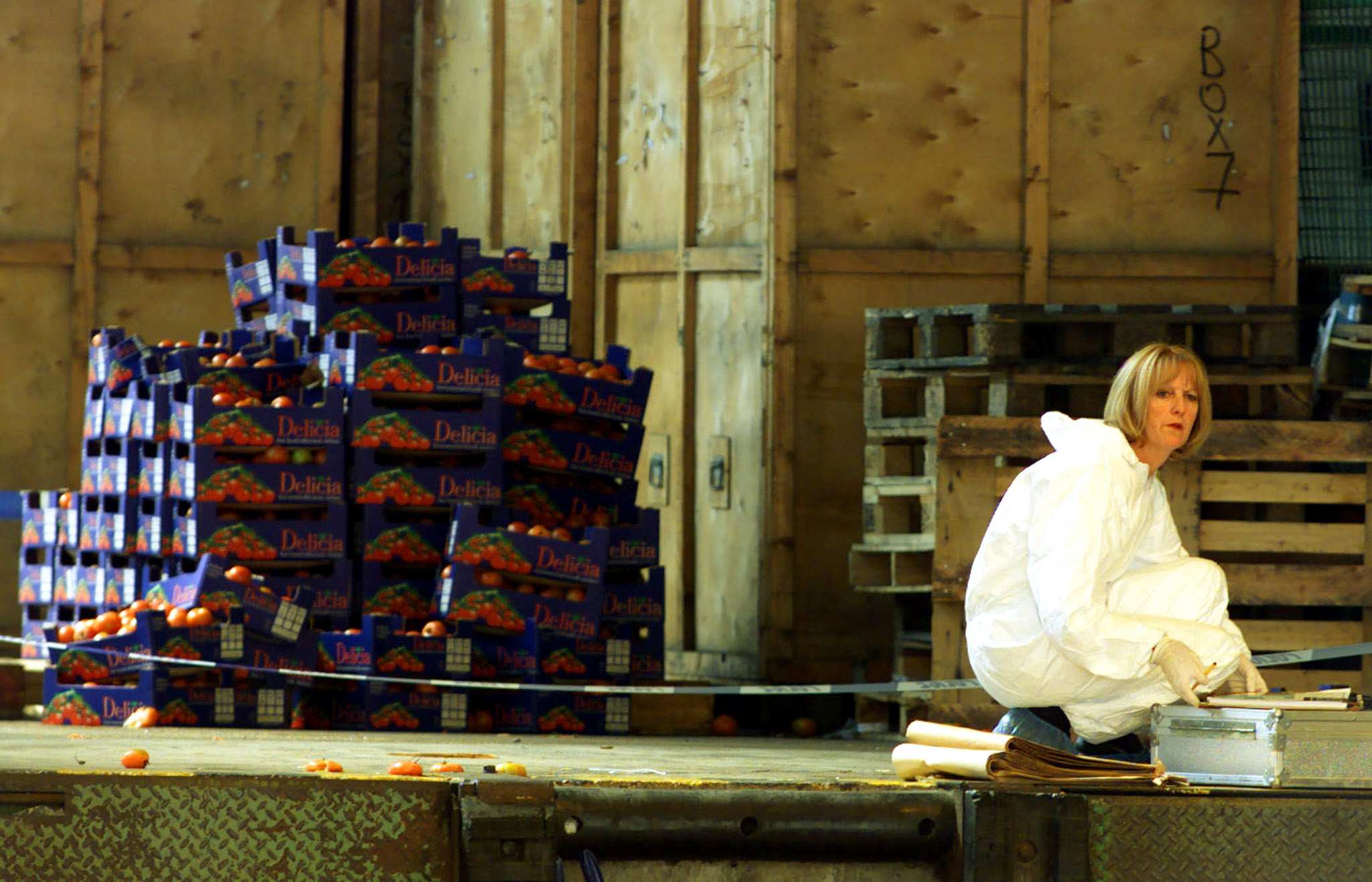 A woman in a white forensic suit crouches on the ground in front of police tape.