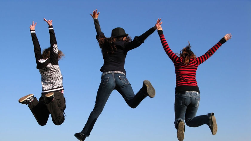 Three girls jump in the air in a grassy field with bright blue sky