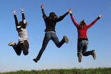 Three girls jump in the air in a grassy field with bright blue sky