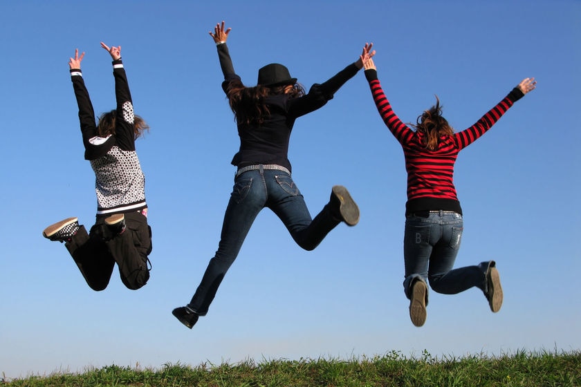 Three girls jump in the air in a grassy field with bright blue sky