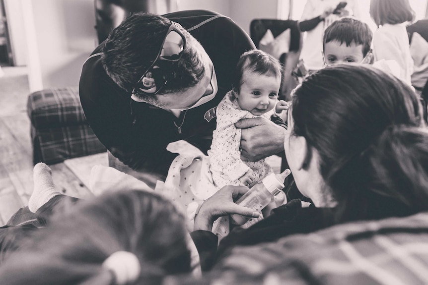 A man wearing a facemask holds his baby daughter, showing the baby to his wife for the first time