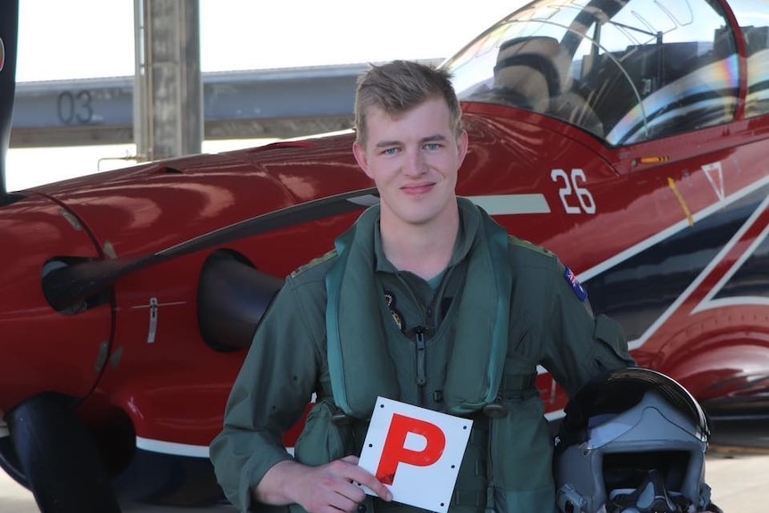 a young man standing in front of a red-coloured light aircraft holding a P Plate