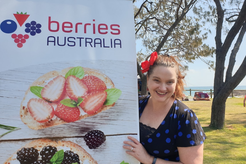 A woman with a pony tail smiles next to a Berries Australia sign with strawberries and blackberries on it.