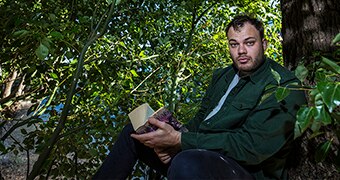 Colour photograph of a man wearing a green shirt sitting in a tree reading a book.