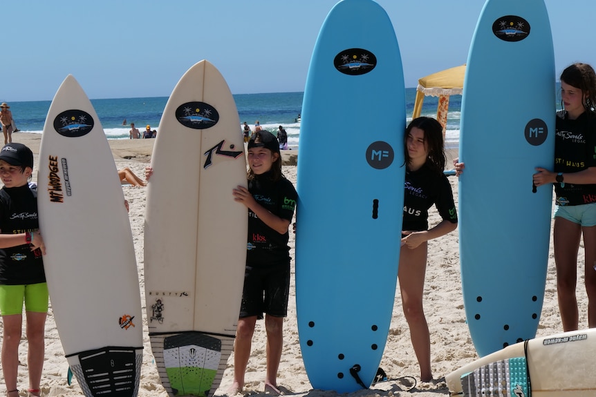 Four children in black rash vests stand in a row holding surfboards.