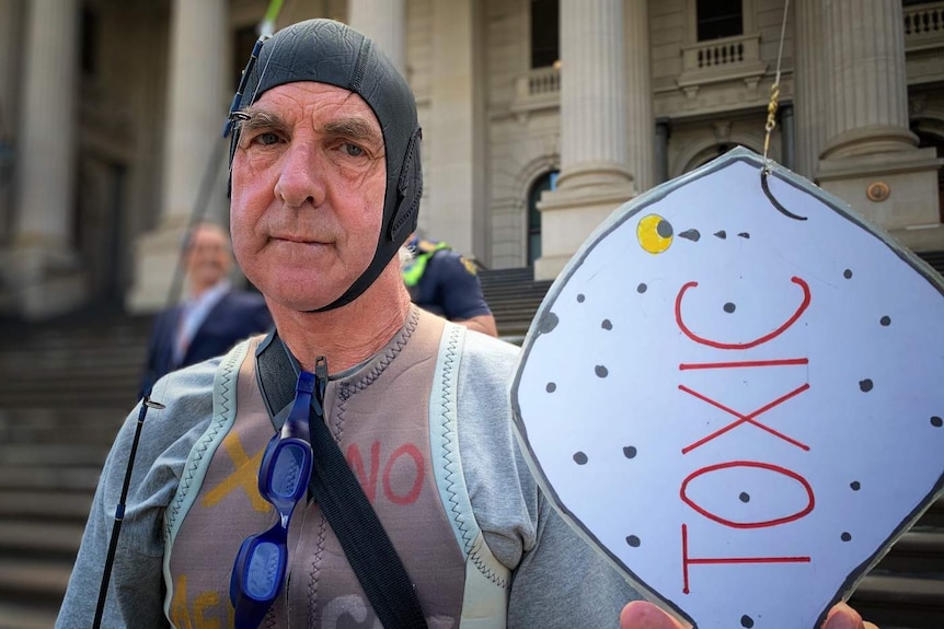 A man wearing a swimming cap and wetsuit holds a cardboard fish labelled 'TOXIC' as he stands outside Parliament House.