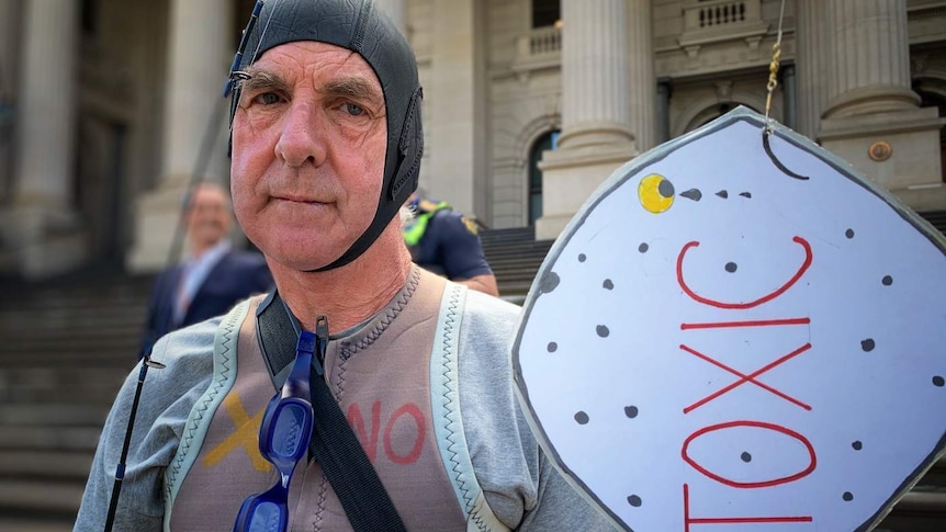 A man wearing a swimming cap and wetsuit holds a cardboard fish labelled 'TOXIC' as he stands outside Parliament House.