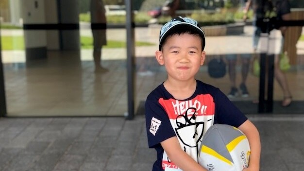 A little boy wearing a cap and holding a ball looks at the camera.