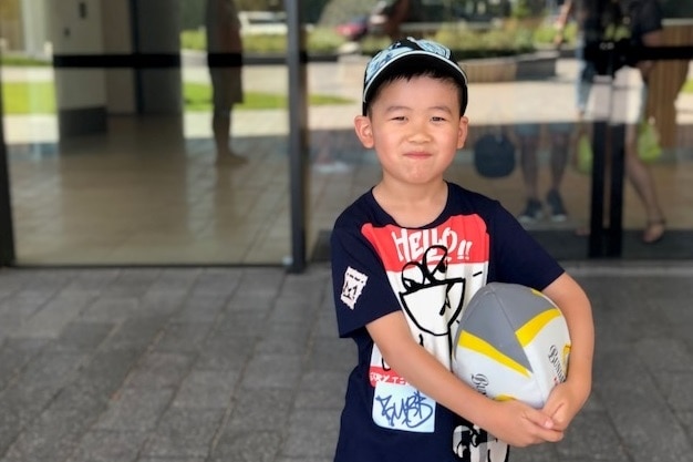 A little boy wearing a cap and holding a ball looks at the camera.