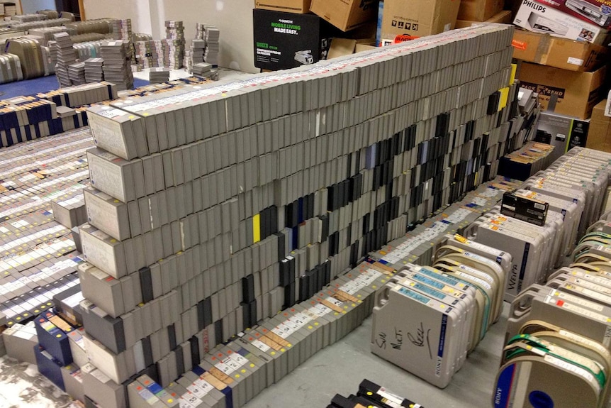 Hundreds of television video tapes in grey boxes stacked in an empty room.