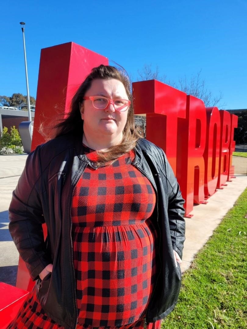 A woman wearing a red and black checked dress and red glasses stands defiantly in front of a red la trobe sign.