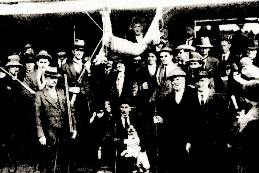 An archival black and white photograph of suited locals with a hanging puma carcus.