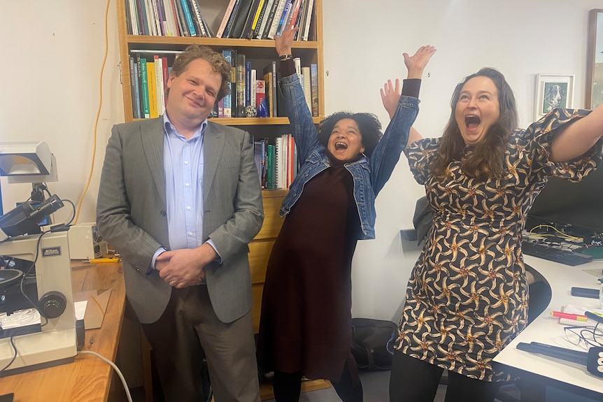 Three people stand, two with arms in arm yelling with joy. 