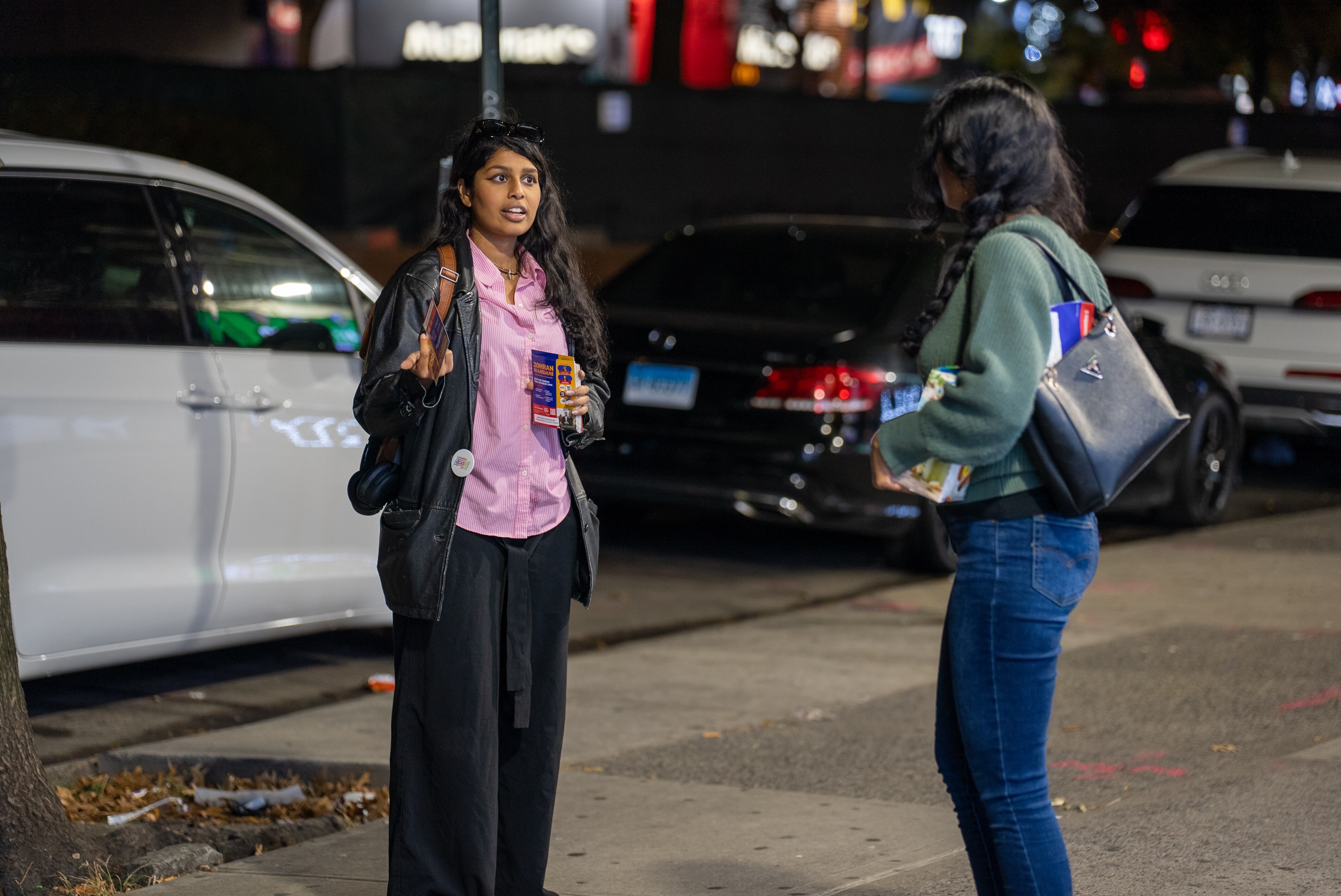 A young woman speaking to another woman on a street at night time.
