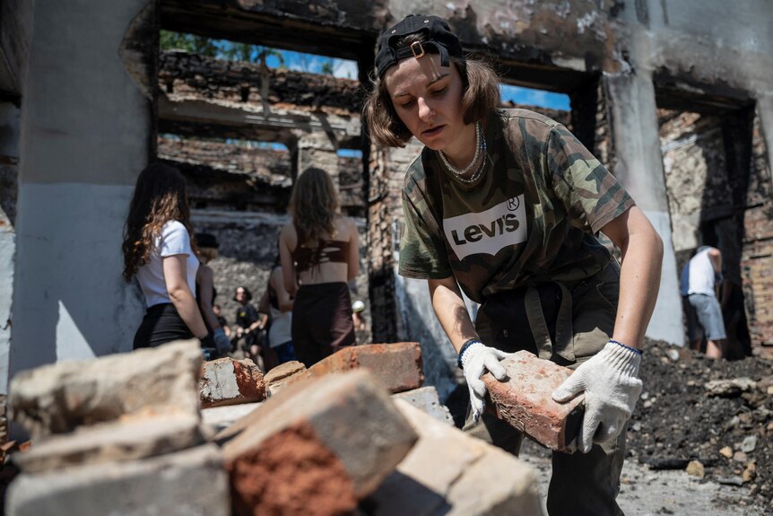 A person in a cap and camo shirt moves bricks in a ruined building