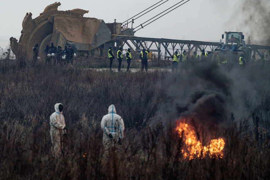 Two people in hazmat suits stand next to a burning pire as police stand guard by an excavator. 