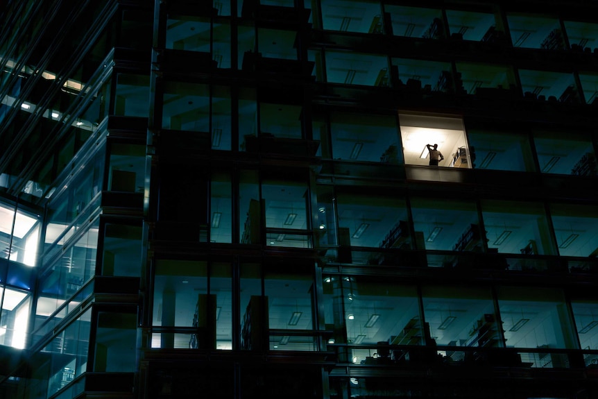 The outside of an office block at night, with just one light on and a person speaking on the phone at the window.
