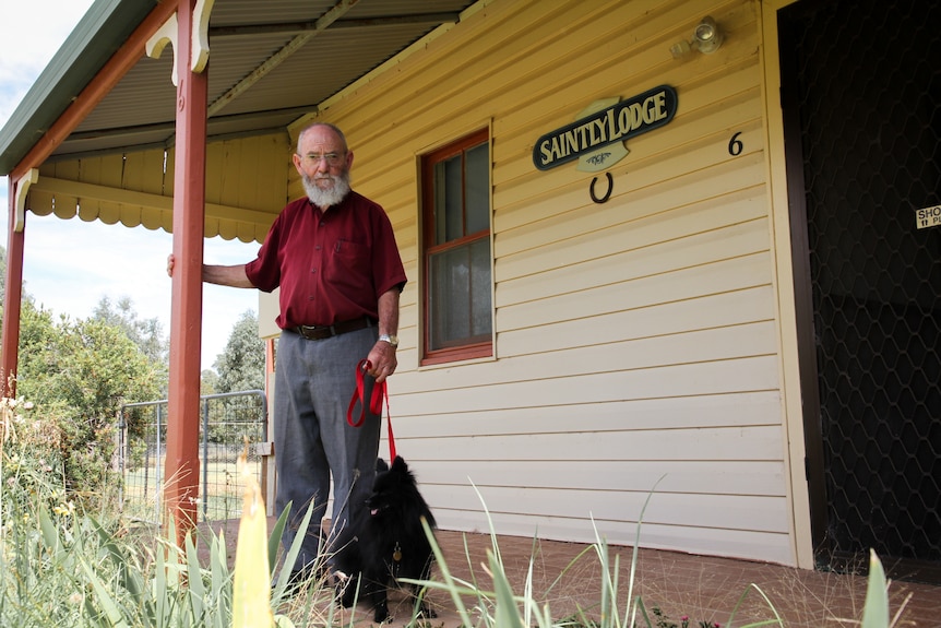 An elderly man with a grey beard stands on the front porch of a home, with his small black puppy on a lead at his feet.
