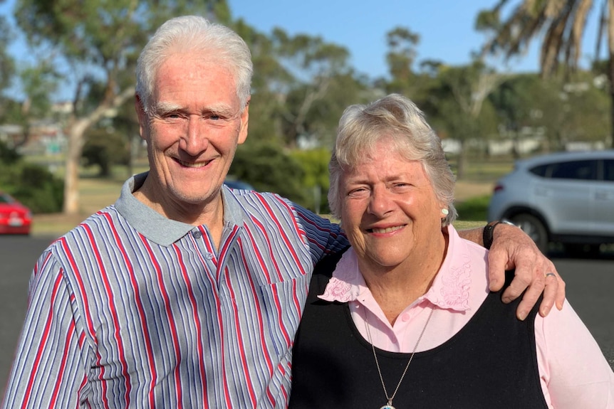 A man and woman stand with arms around each other as they stand in the street in front of gum trees.