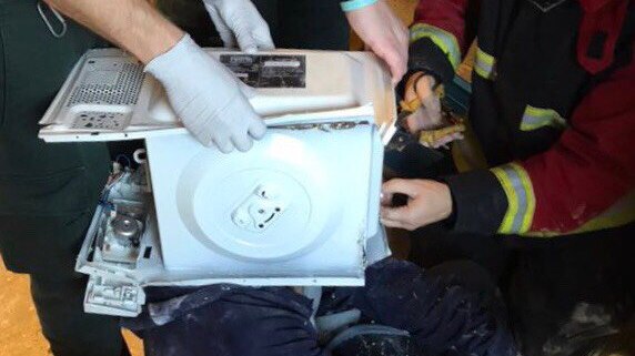 West Midlands fire authorities try and remove a man's head from inside a microwave oven. He sits on the ground cross-legged.