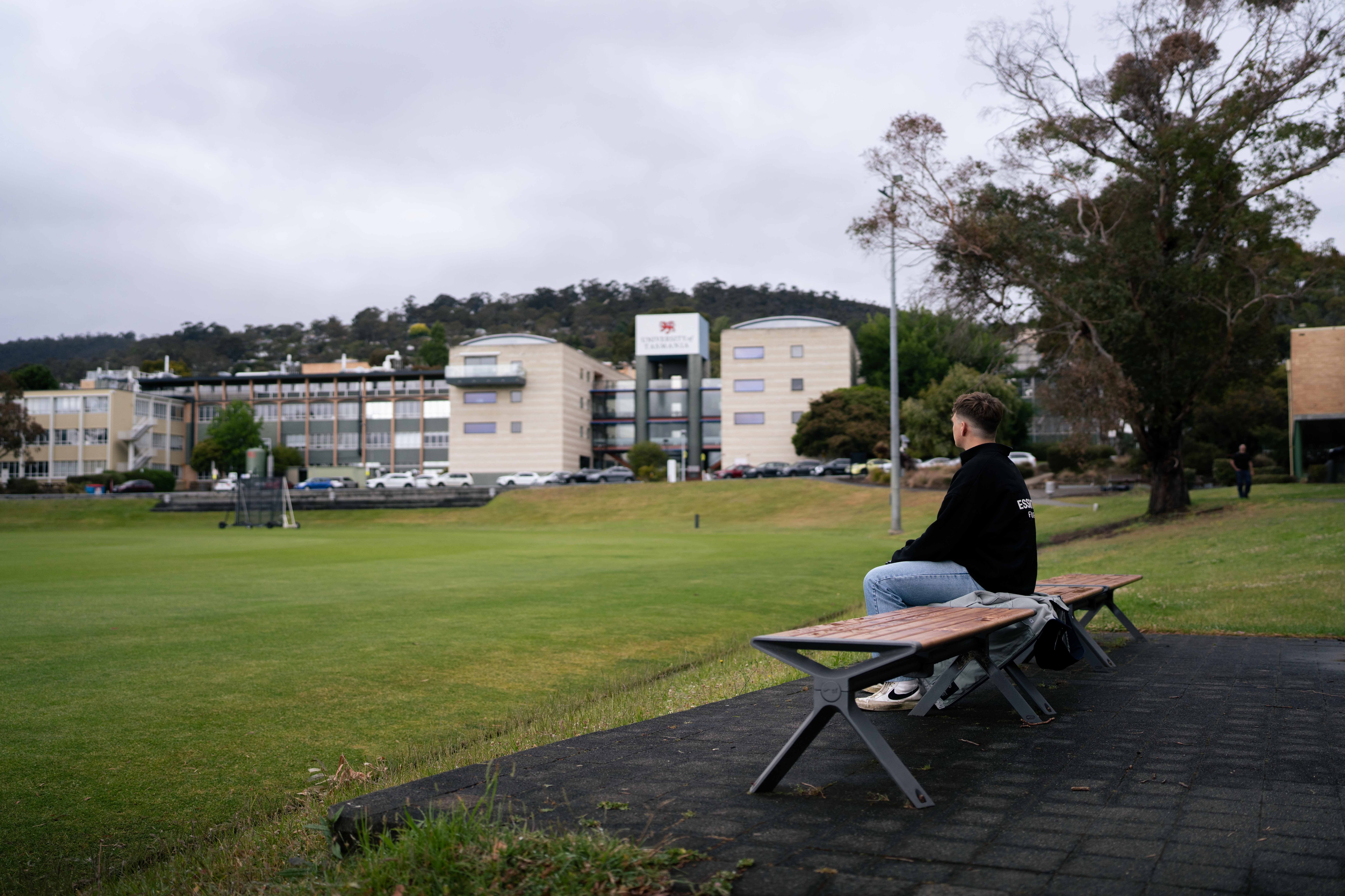 Man sits on park bench looking out to a university in the distance