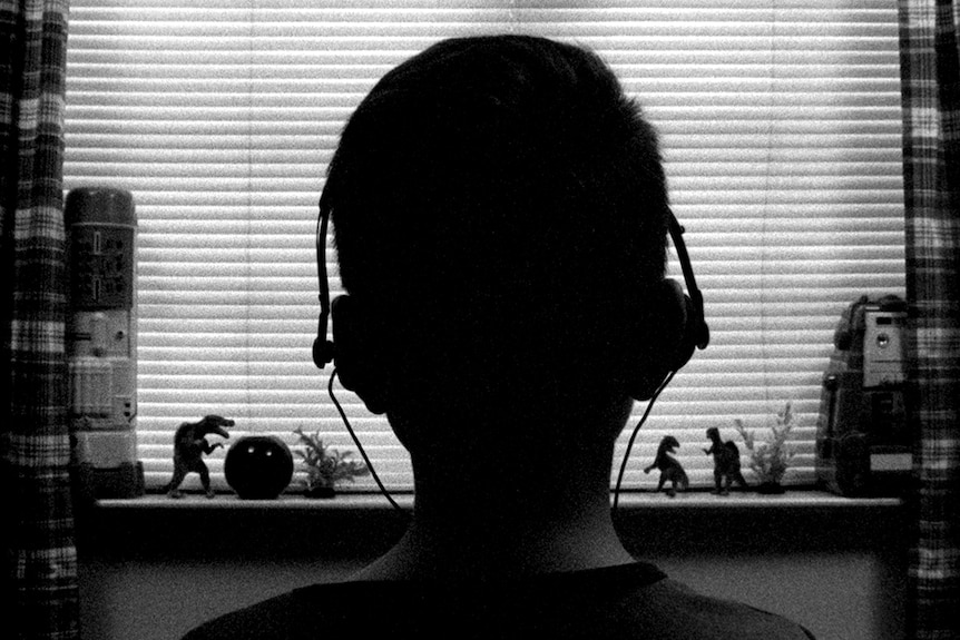 Black and white still of Aidan Langford wearing headphones with back to camera in front of bedroom window in 2018 film 1985.
