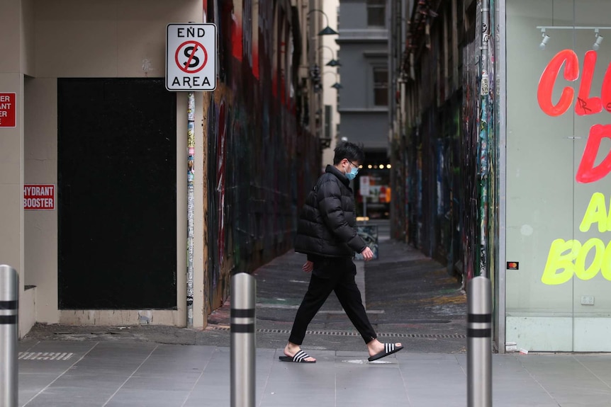 Man walks past a Melbourne alley. He is wearing a puffy jacket and a mask. A sign near him reads 'closing down'.