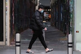 Man walks past a Melbourne alley. He is wearing a puffy jacket and a mask. A sign near him reads 'closing down'.