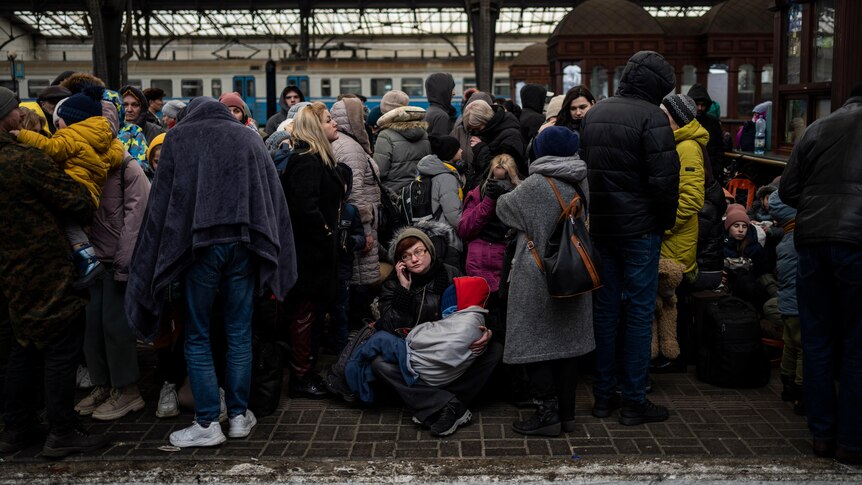 A train station platform, with people wearing blankets and winter clothes all huddled.
