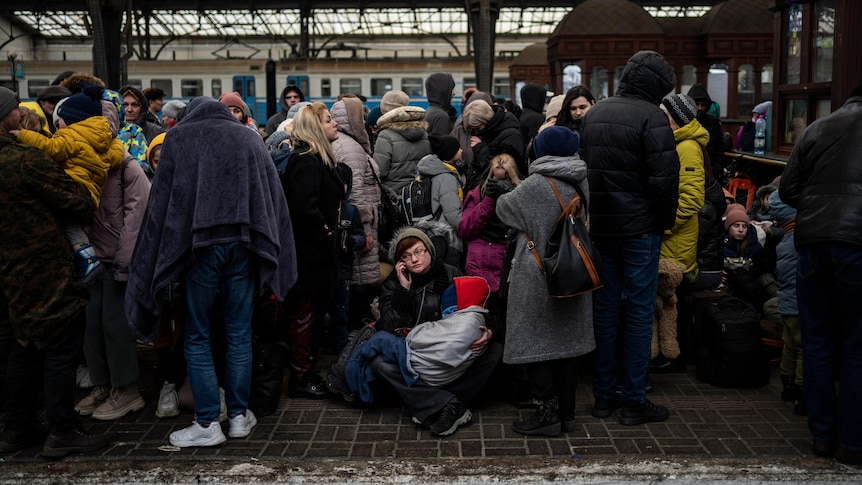 A train station platform, with people wearing blankets and winter clothes all huddled.