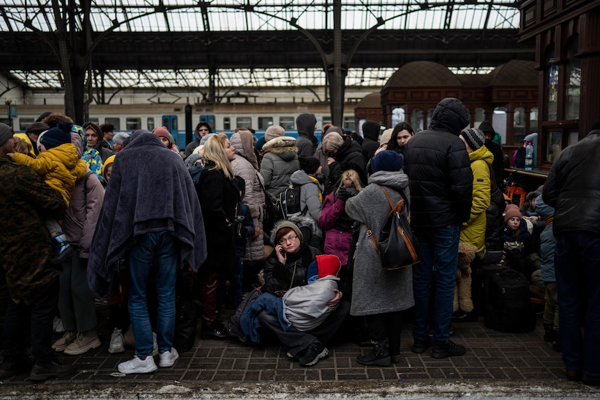 A train station platform, with people wearing blankets and winter clothes all huddled.