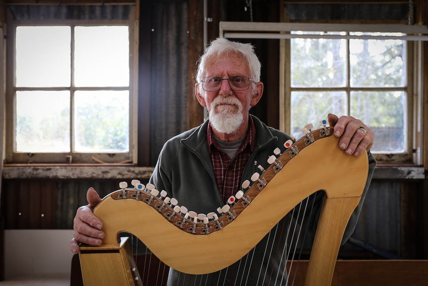 An older man stands behind a harp in a workshop-shed space.
