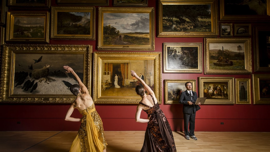 People stretch in front of paintings in gold frames hung on a red wall in a gallery.