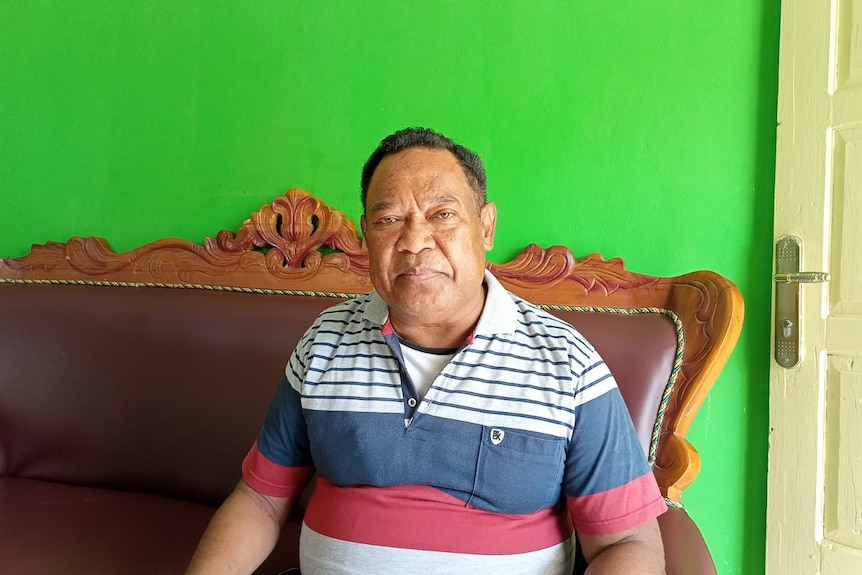 An Indonesian man in a polo shirt sits on a couch, looking serious 