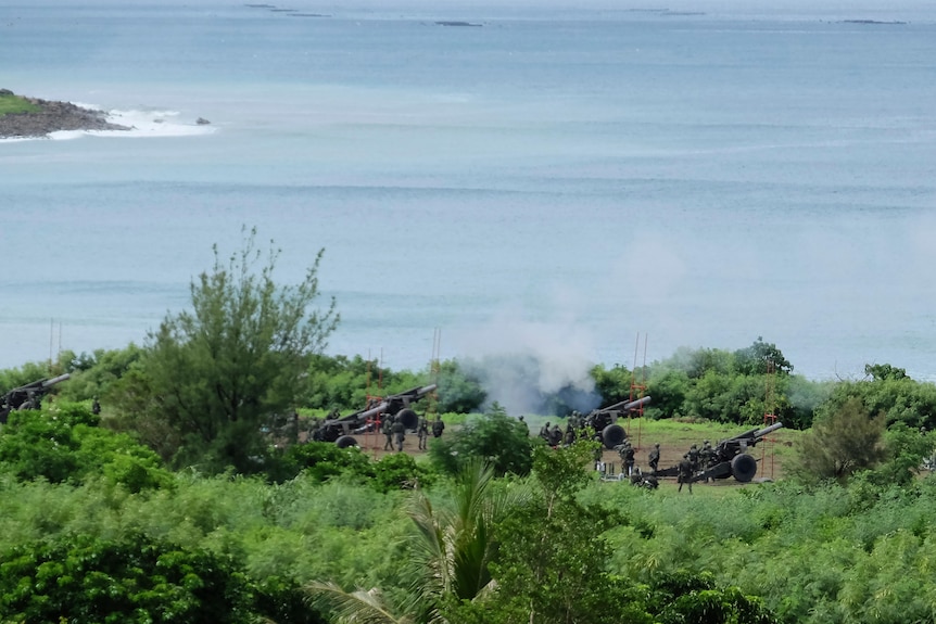 Three artillery crews fire live rounds out to sea from an elevated clearing.