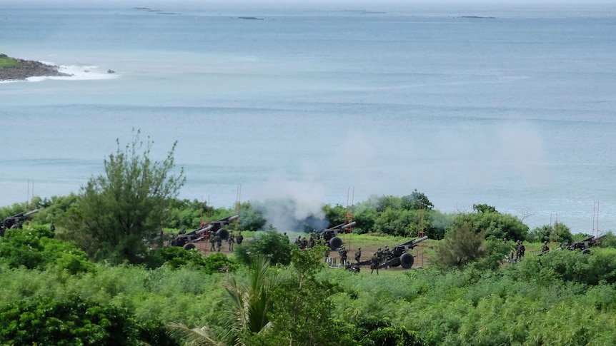 Three artillery crews fire live rounds out to sea from an elevated clearing.
