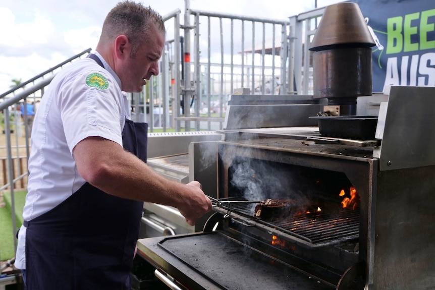 Chef Sam Burke using tongs to flip a steak, hot coals and smoke coming from grill.