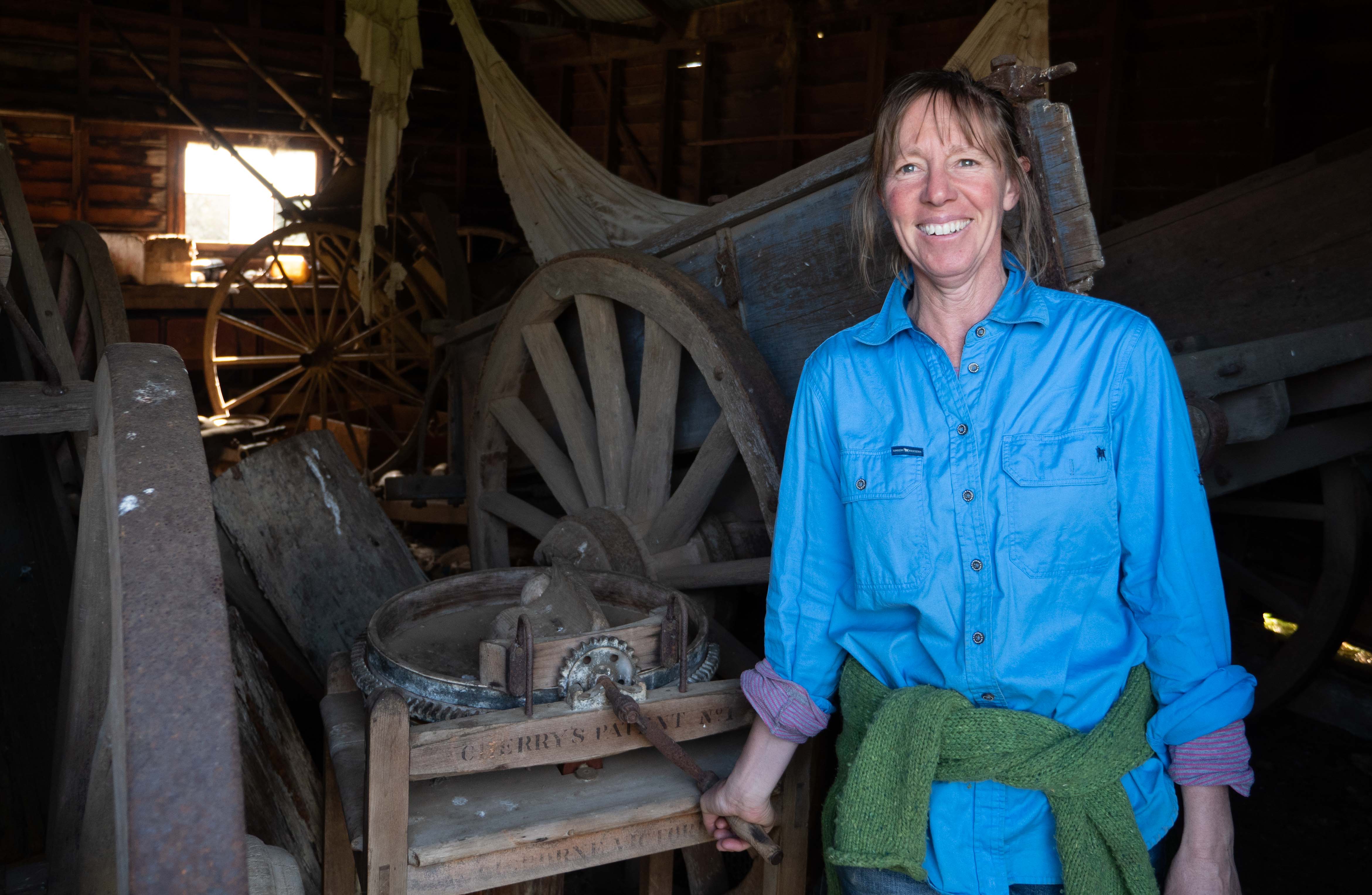 A woman in a blue shirt standing in an old shed, with old wagon wheels behind her
