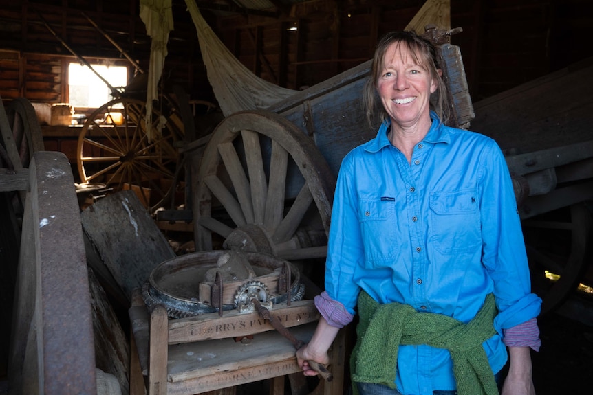 A woman in a blue shirt standing in an old shed, with old wagon wheels behind her
